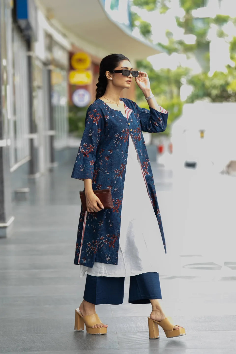 A woman walking outdoors wearing a navy blue floral kurta over a white tunic, navy cropped pants, beige platform sandals, and holding a brown clutch.