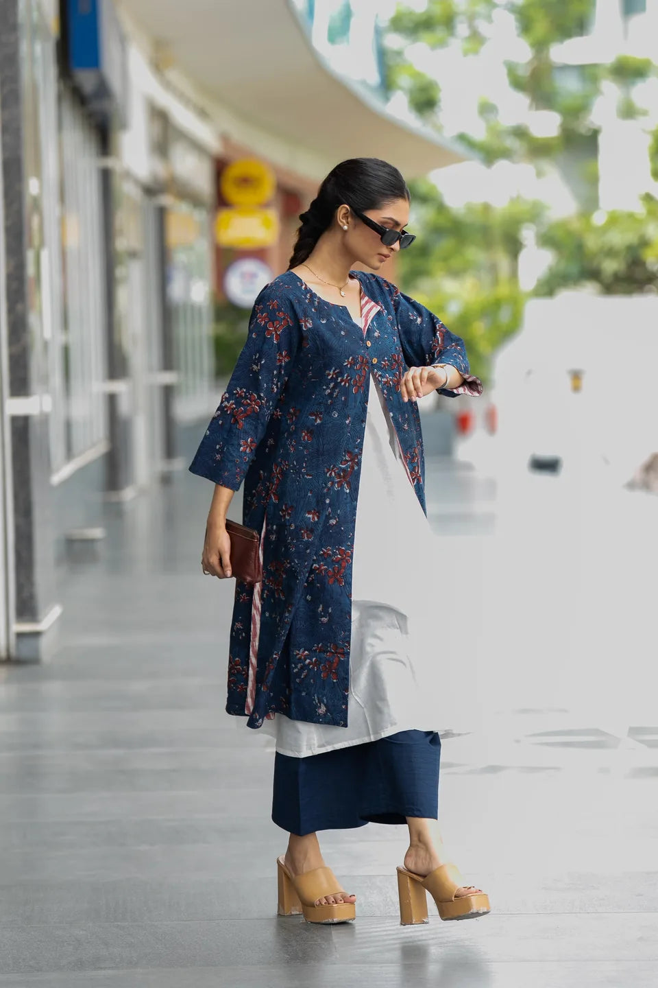 A woman walking outdoors wearing a navy blue floral kurta over a white tunic, navy wide-leg pants, and beige platform sandals, with sunglasses and a small brown clutch.