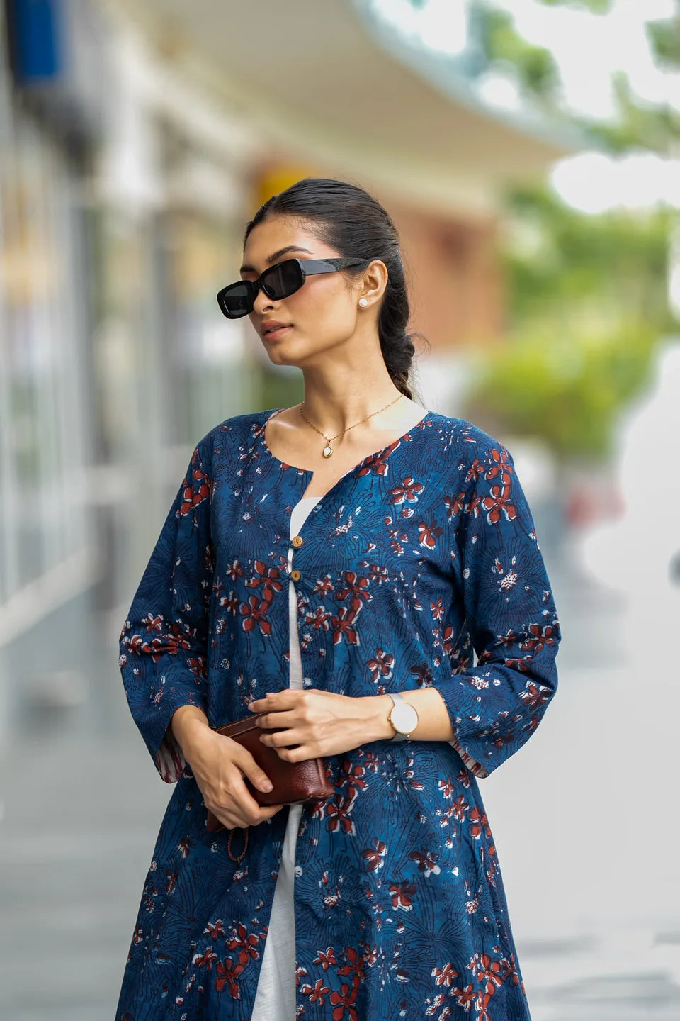 A woman wearing a navy blue floral tunic with red and white patterns, styled with black sunglasses, a gold necklace, and a white undershirt, standing in an outdoor setting.