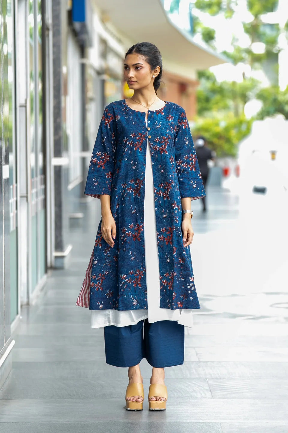A woman modeling a three-piece navy blue kurta set with a red and white floral print, featuring a long tunic, matching wide-leg pants, and a white underskirt, set against a blurred outdoor walkway.