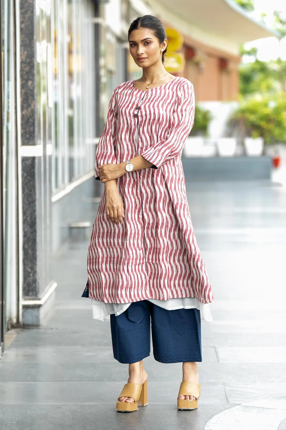 A woman modeling a pink and white vertically striped kurta with three-quarter sleeves, paired with navy blue cropped pants and tan platform sandals, standing in an outdoor walkway.